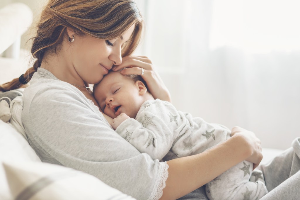 Dark haired mom with hair in braid holding newborn baby against her chest. Baby is asleep after breastfeeding. Mom is gently smiling. She has one hand on baby's body and one hand on his head. Her face is touchung baby's forehead. Mom is bonding with her newborn.