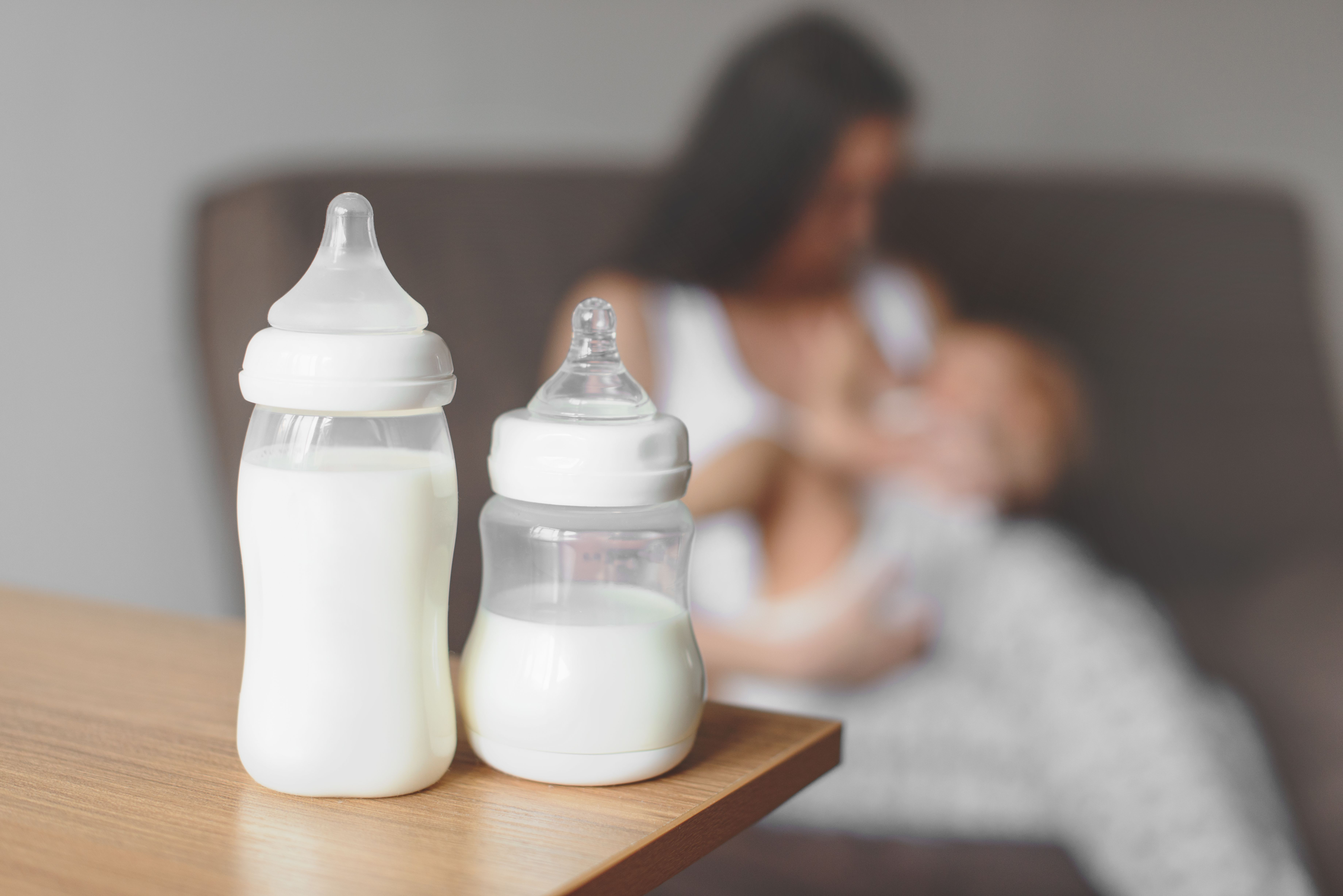 Bottle-feeding baby 
Two bottles full of milk sitting on table. In background is blurred image of mother cradling baby in her arms 