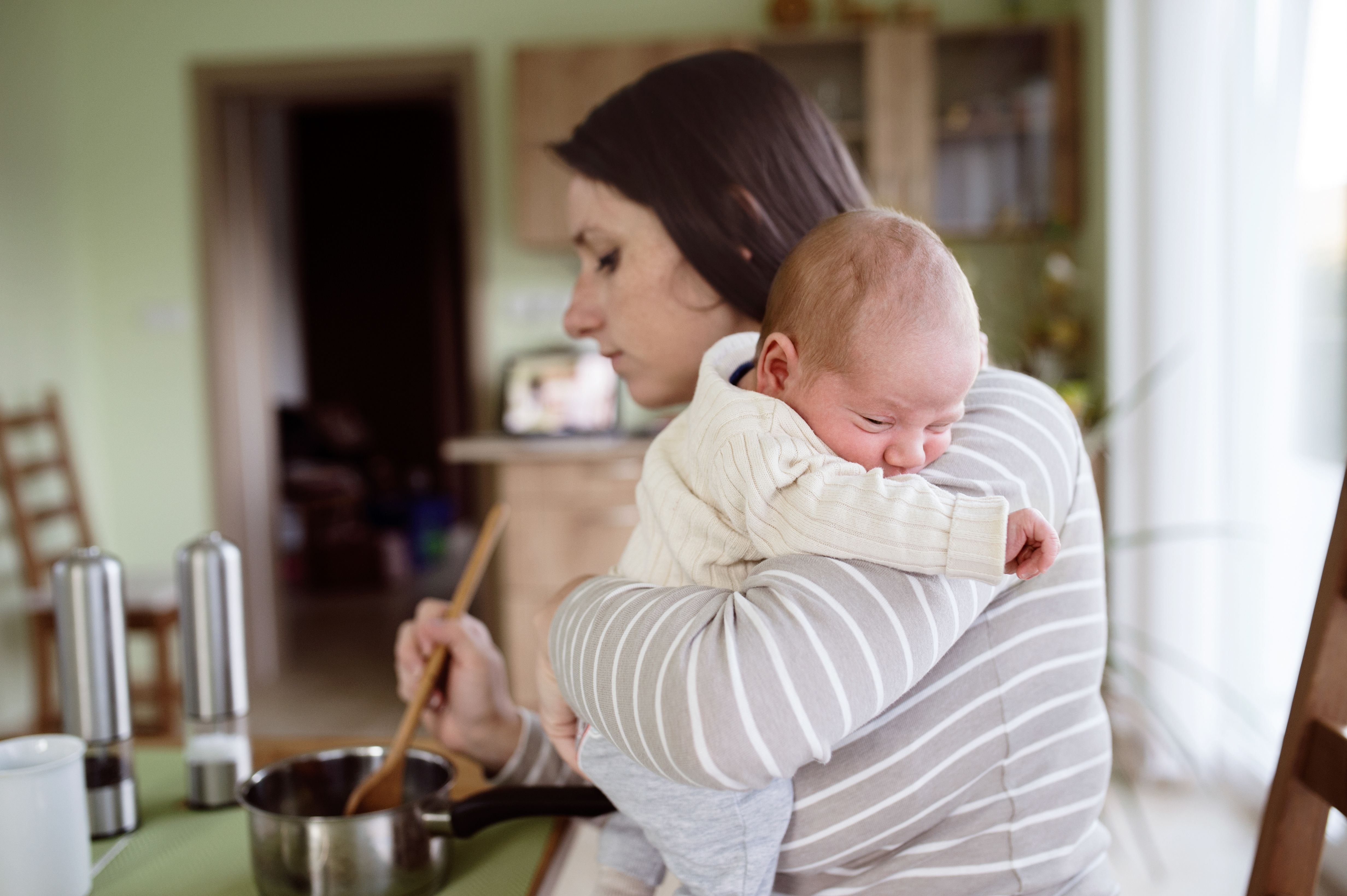 Mother is in the kitchen, holding her sleepy newborn baby on her shoulder. With her other hand she is holding a wooden spoon and stirring food in a pot. Mother is cooking dinner as she holds and comforts her baby.