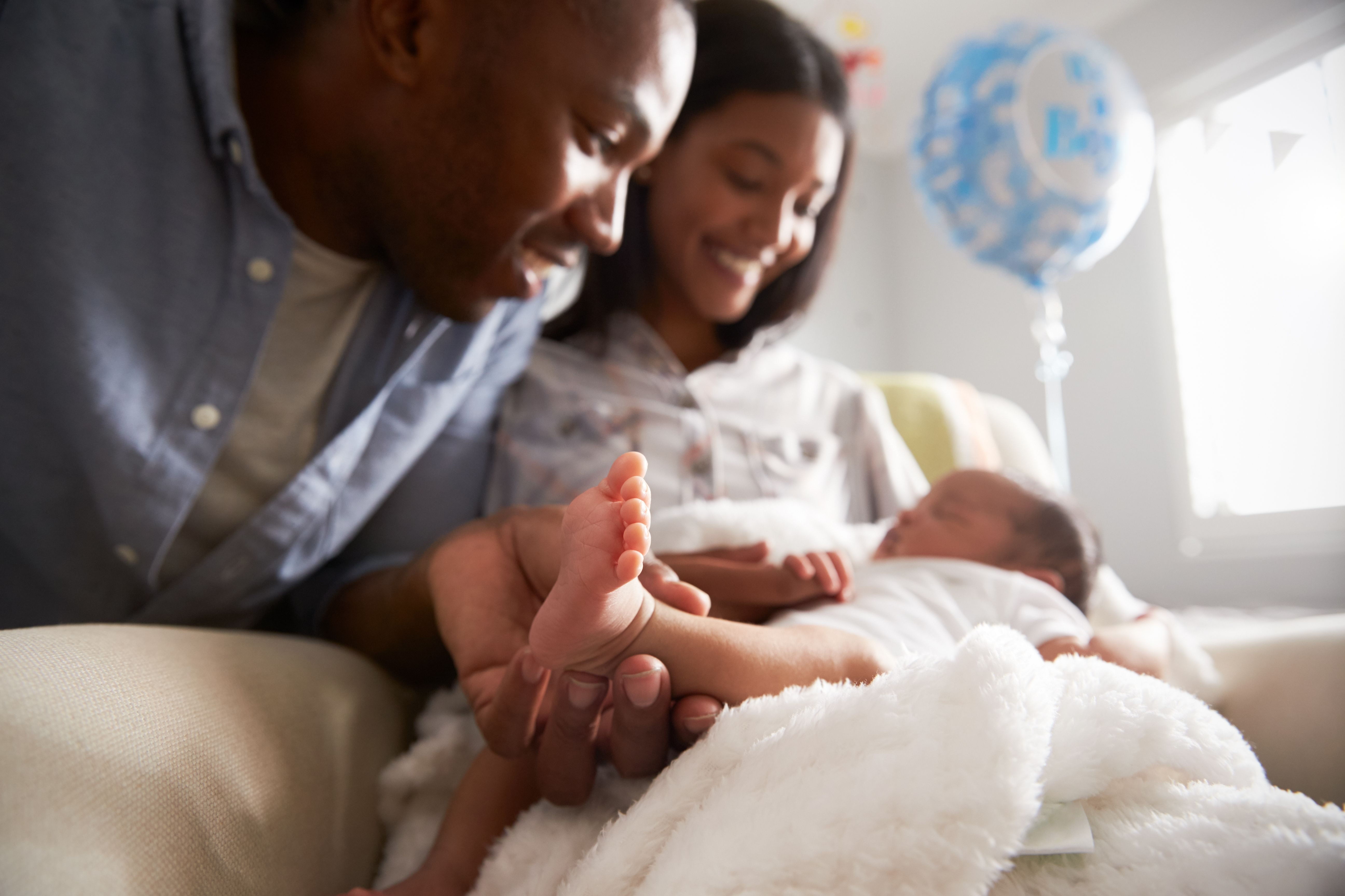 Black mother and father sitting in comfortable chair happily looking at new baby who is wrapped in soft blanket. Dad is holding baby's foot and mom is holding baby's hand. They look overjoyed to be home with their new baby. Balloon in background shows that family is celebrating baby's birth. 