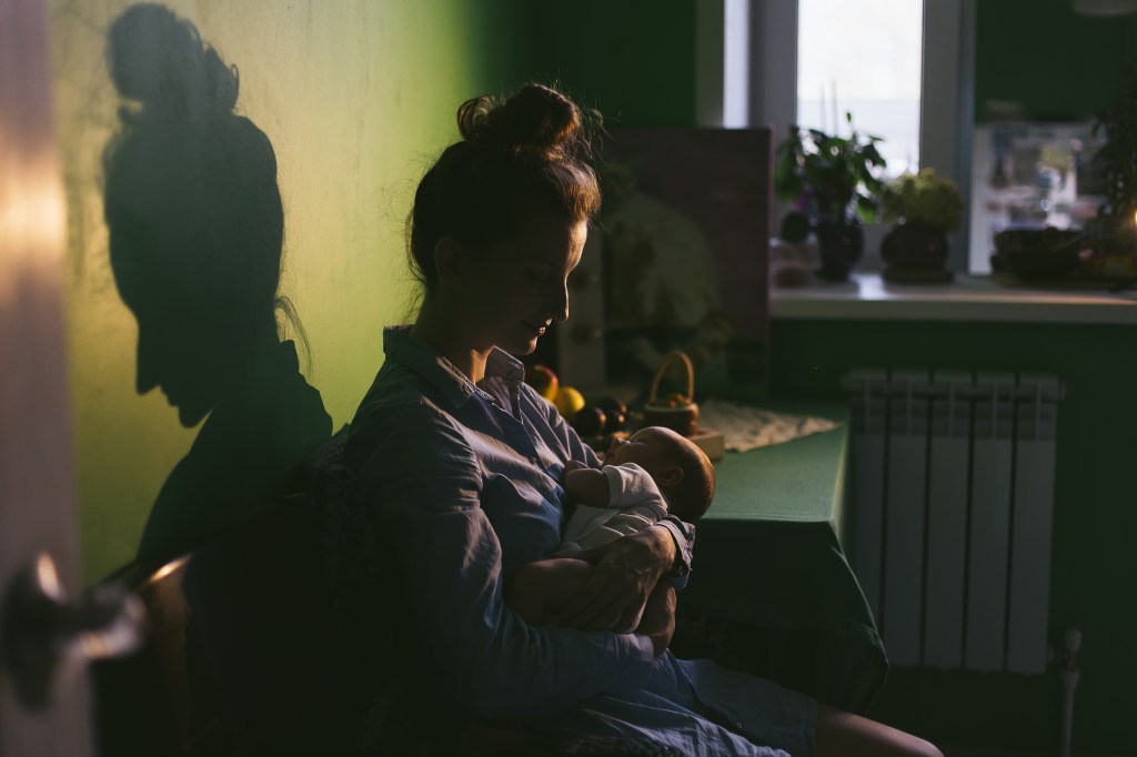 Mother sitting in dimly- lit room holding infant
Her hair is in a messy bun and she is wearing a night shirt
Mother looks tired but peaceful as she holds sleeping baby
Waking up with baby in the middle of the night