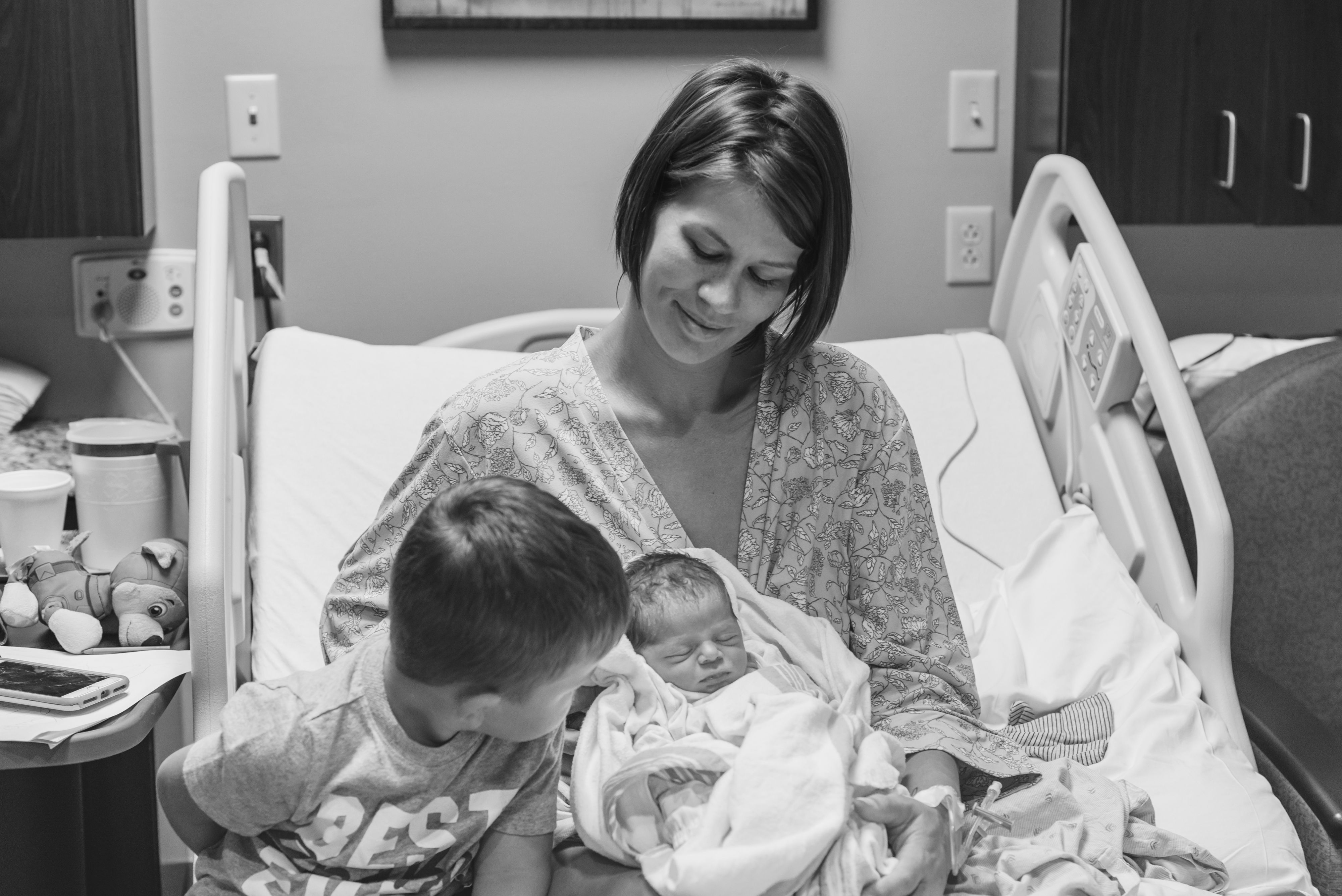 Mother in hospital bed with newborn baby. She is smiling as she holds the baby and his sibling is meeting his new brother/ sister.